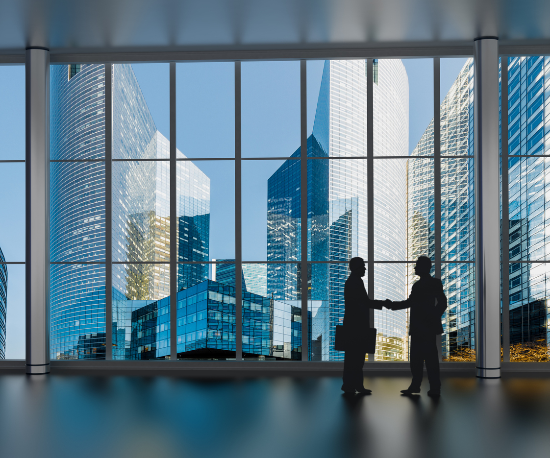 from a far two people, one holding a briefcase, shake hands in front of a large wall of glass windows showing tall city building