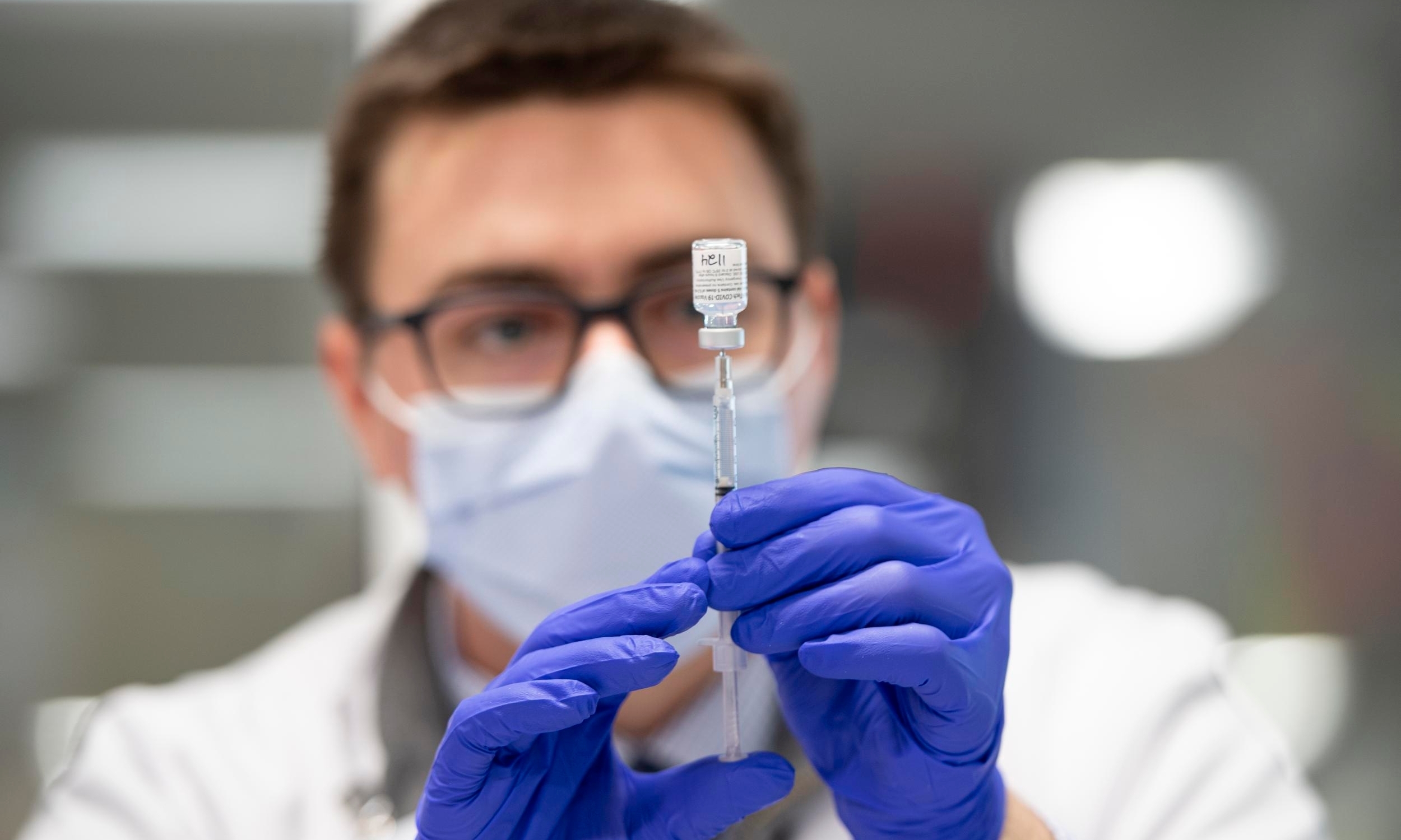 A mean wearing a mask and gloves holds up a medical tube to look at the contents closely